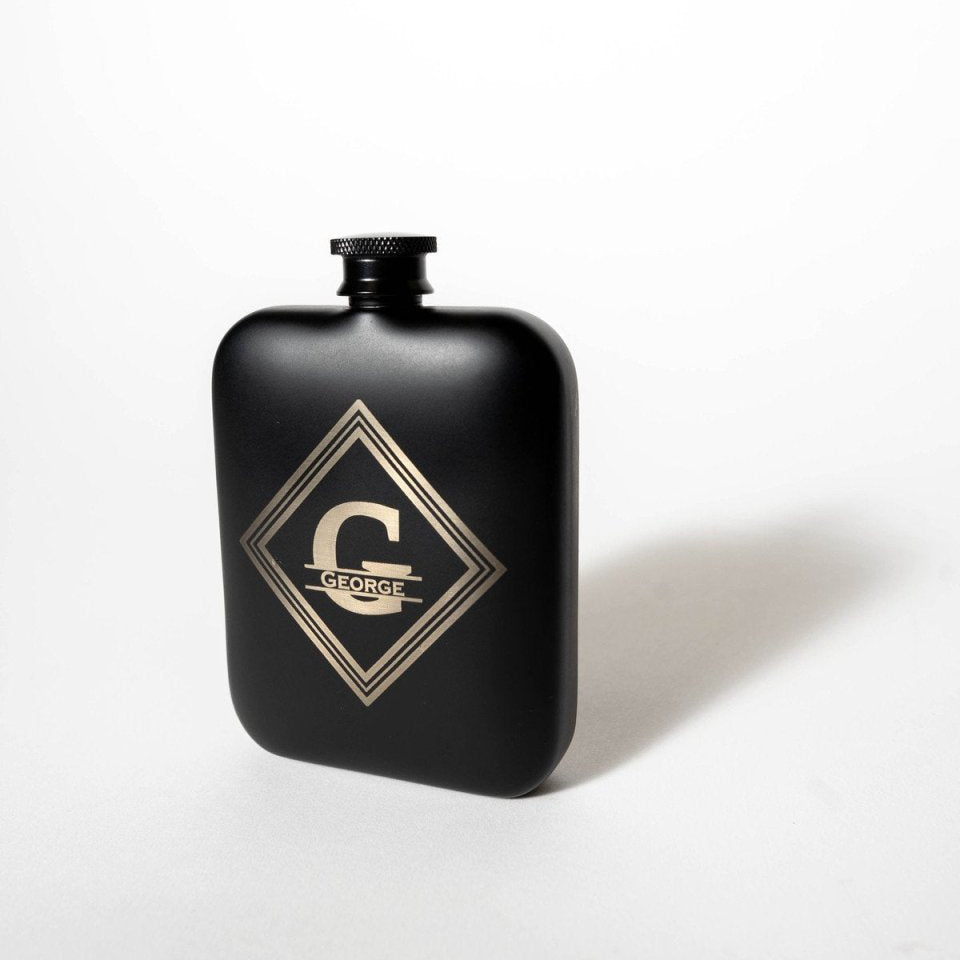 Matte black groomsmen flask standing on a white background, engraved in silver with a large G, the name GEORGE and a diamond border