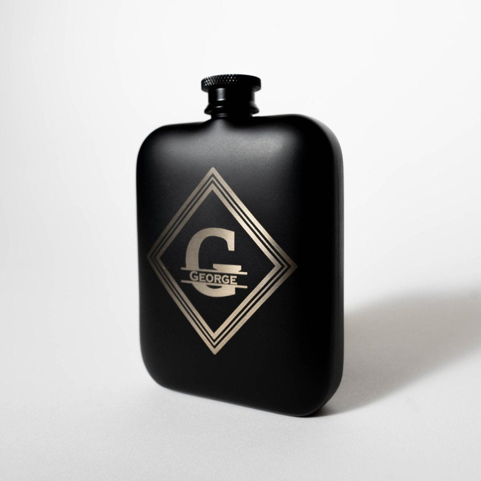 Angled view of the matte black Hip To Sip flask with its knurled screw cap and the engraved G and GEORGE monogram inside a triple-line diamond