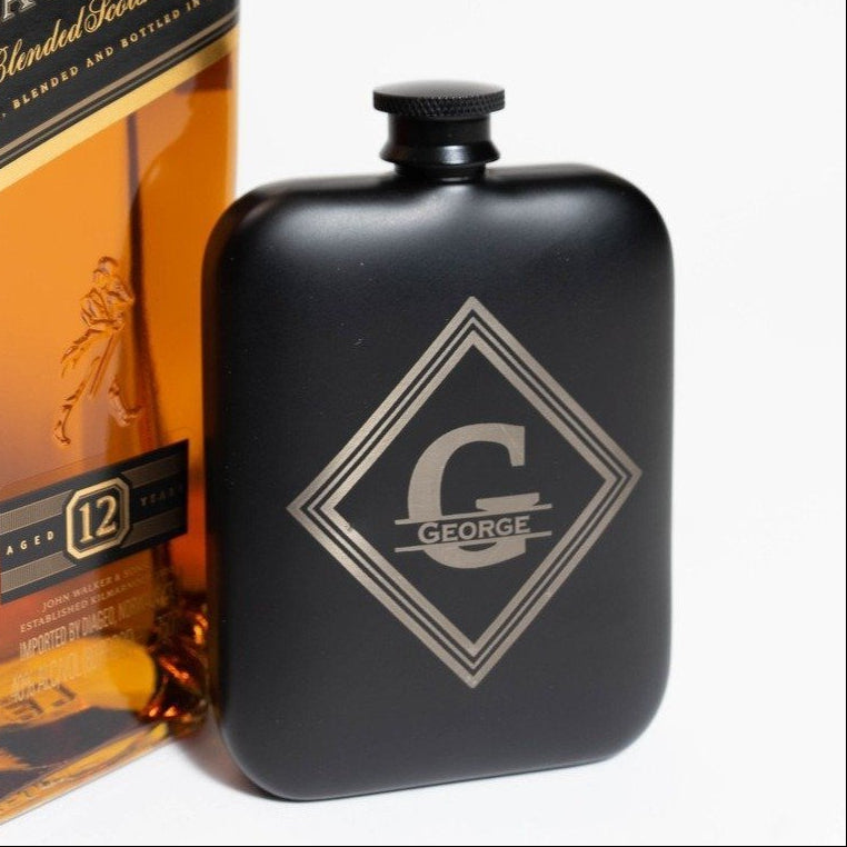 Personalized 6 oz black flask engraved GEORGE across a big letter G inside a diamond, standing beside a bottle of whisky to show its pocket size