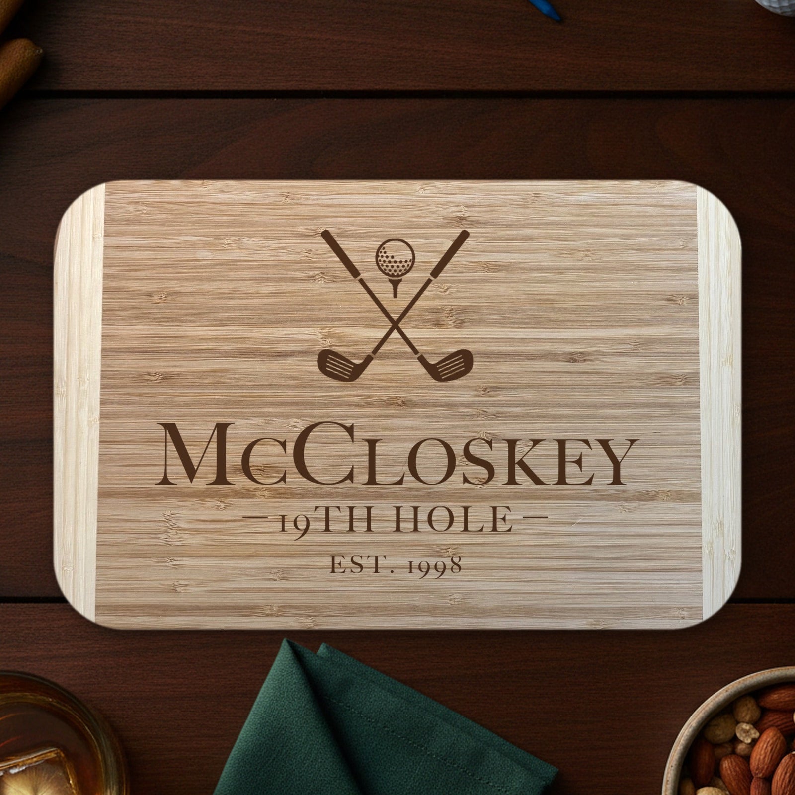 Personalized golf groomsmen cutting board on a dark walnut bar top with golf balls, tees and whiskey, engraved McCloskey 19TH HOLE EST. 1998 under crossed clubs.