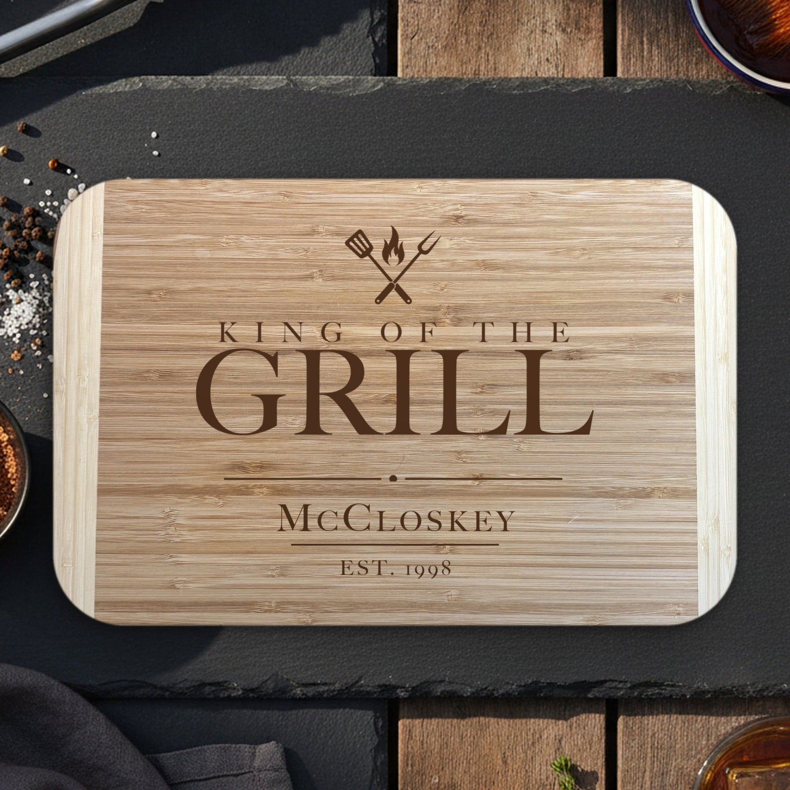 King of the Grill cutting board for dad on dark slate with grilling tongs, dry rub and bourbon, engraved McCloskey EST. 1998 under crossed grill tools on two tone bamboo.