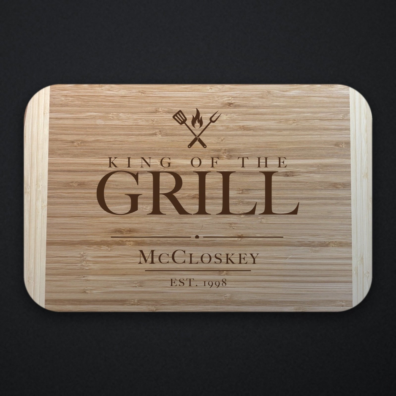 Father of the groom King of the Grill cutting board in two tone bamboo engraved under crossed grilling tools and a flame, with McCloskey and EST. 1998 below.