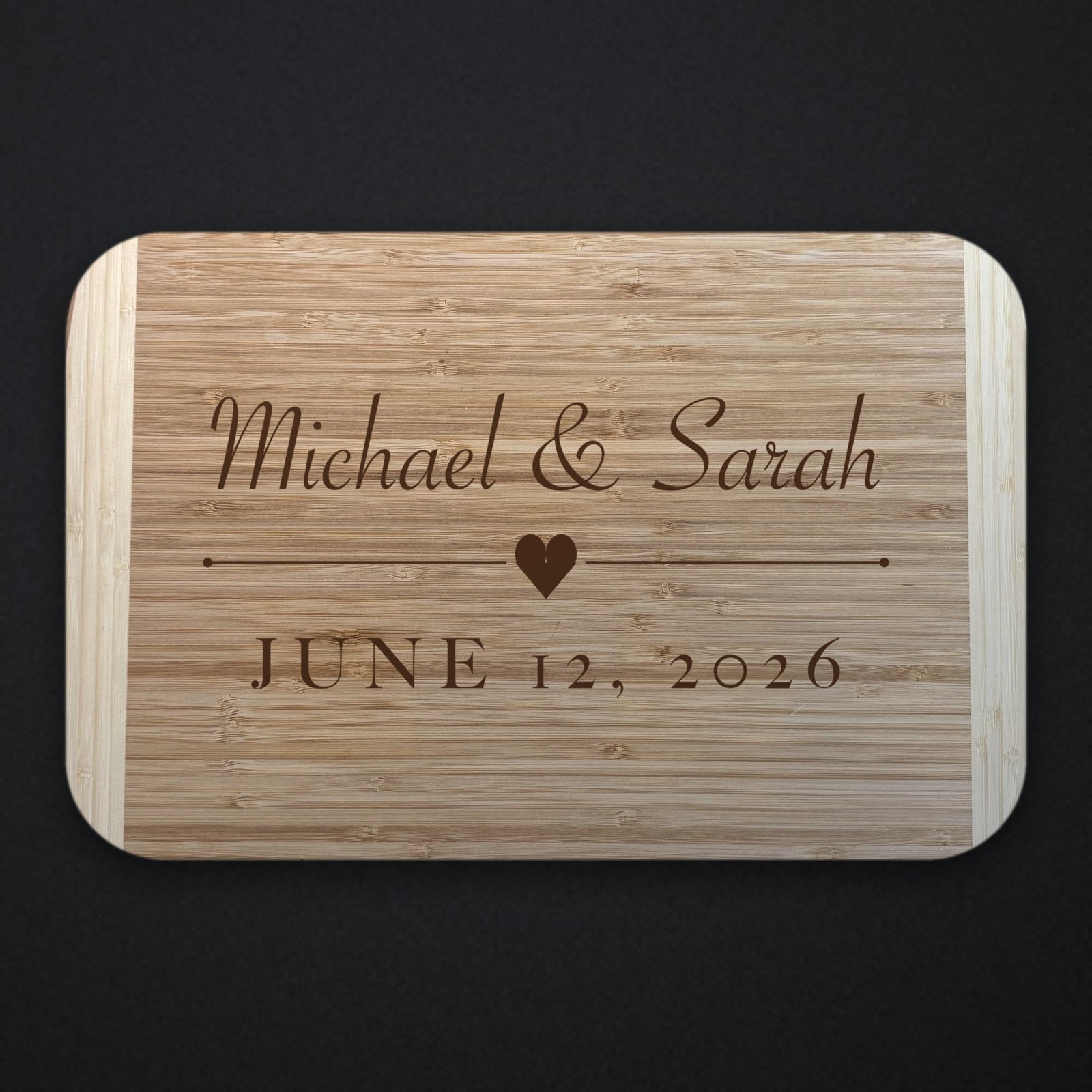 Two tone bamboo wedding cutting board engraved with Michael and Sarah in script above a heart and rule, with JUNE 12 2026 beneath, shown on a dark background.