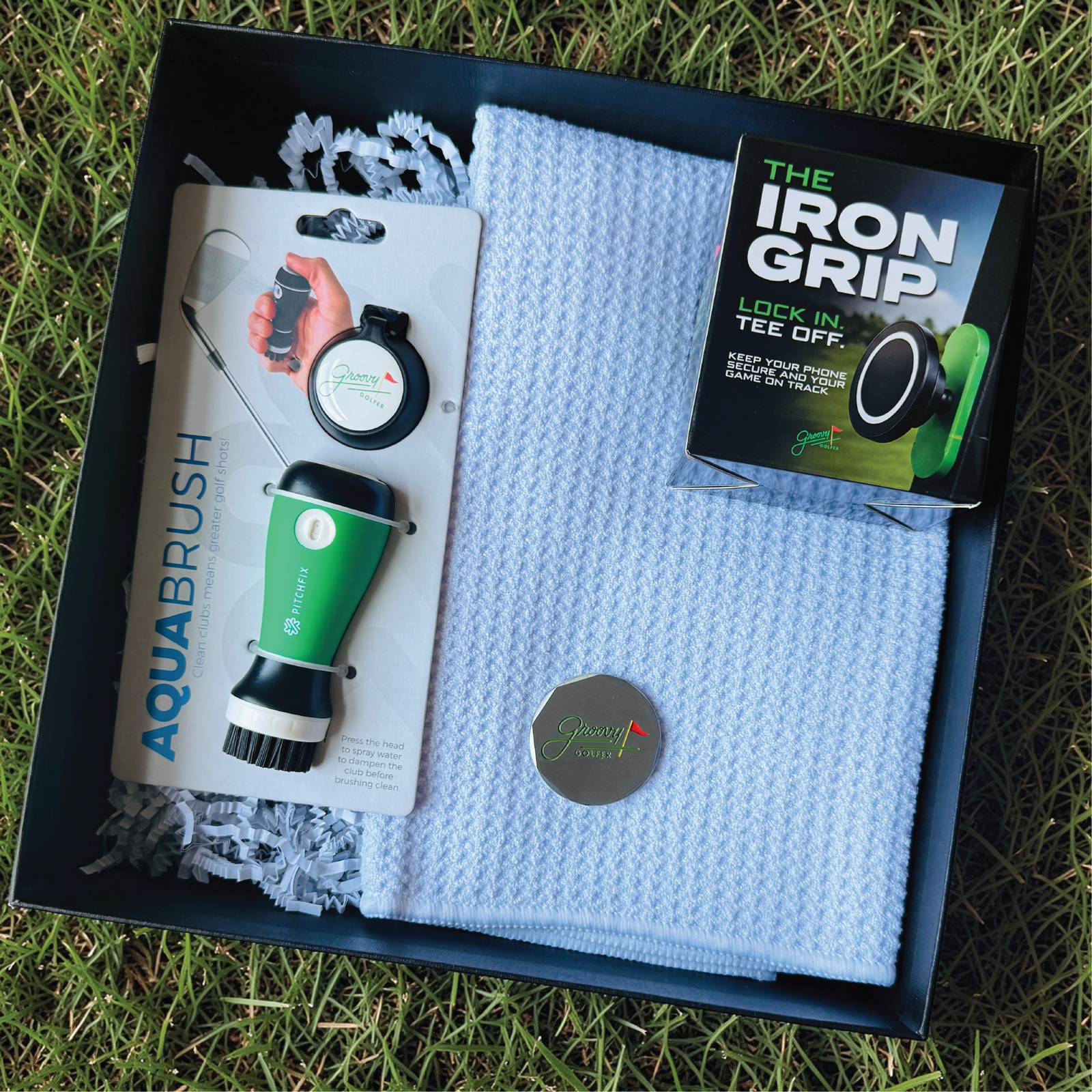 Flat lay of the open groomsman golf gift set showing the AquaBrush club cleaner, blue waffle towel, magnetic phone mount and golf ball marker.