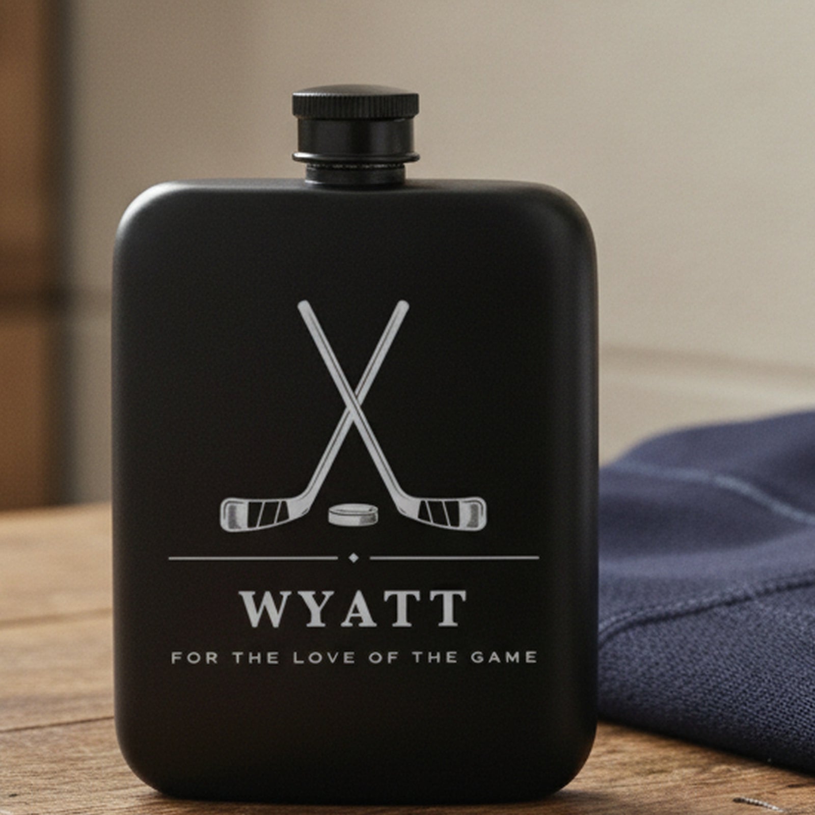 Engraved hockey flask reading For The Love Of The Game on a locker room bench beside a folded navy jersey and a rolled towel.