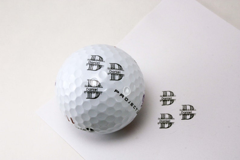 White golf ball with sample personalization showing "Daniel" name and monogram stamp design, displayed next to individual stamp impressions on white background