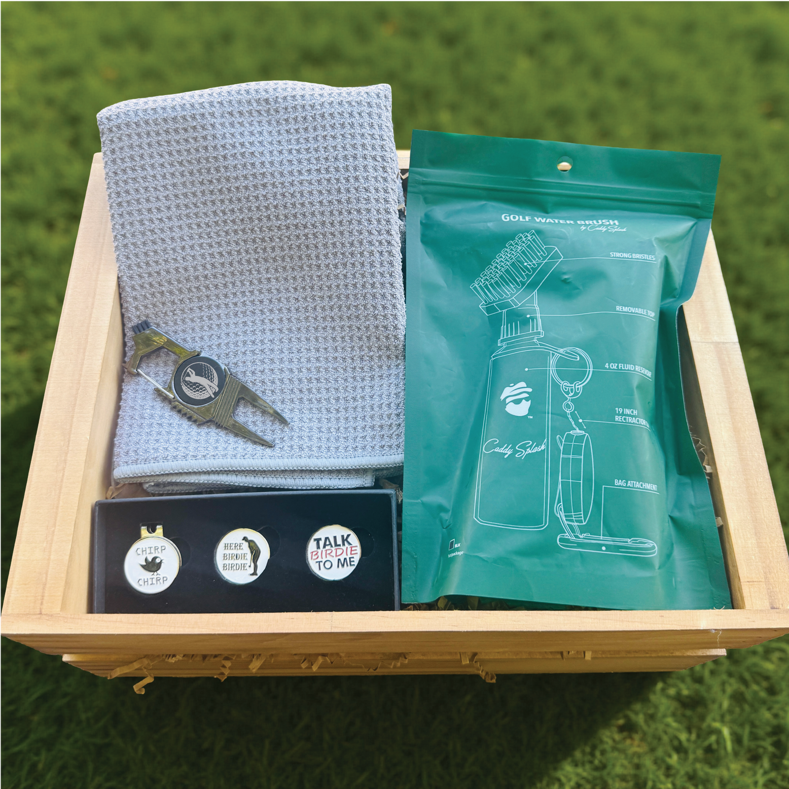 Golf gift set in wooden crate containing gray textured towel with divot tool, three golf ball markers with phrases, and turquoise water brush package on grass background