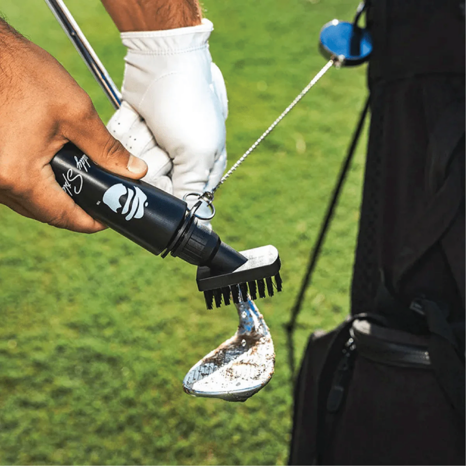 Black golf club brush with personalized monogram design being used on golf shoe on green course, held in golfer's hand wearing white glove