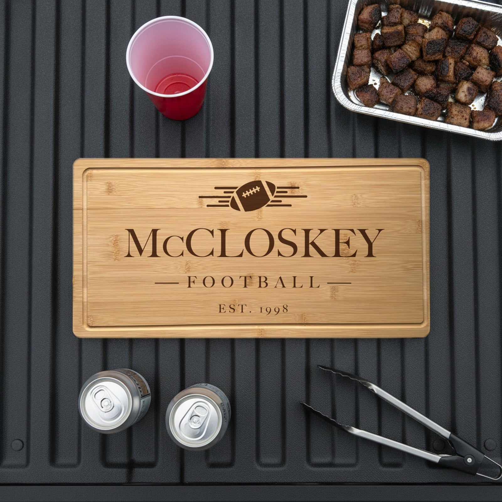 Engraved football bamboo board on the open tailgate of a pickup truck beside a tray of grilled burnt ends, two cans, a red cup and grill tongs.