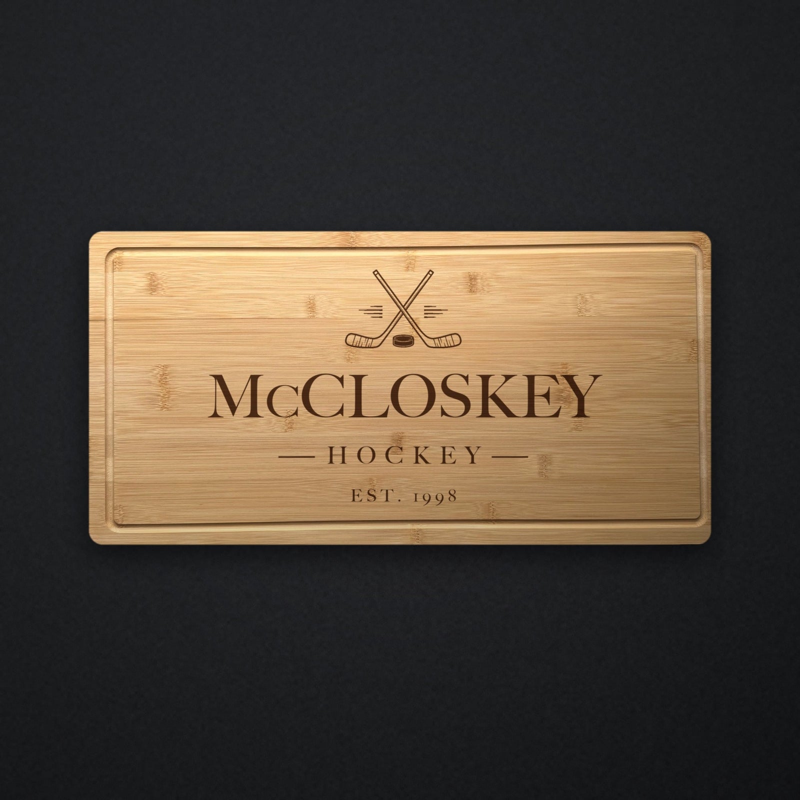 Personalized hockey cutting board in solid bamboo on dark slate, engraved with crossed hockey sticks and a puck above the family name and the year.