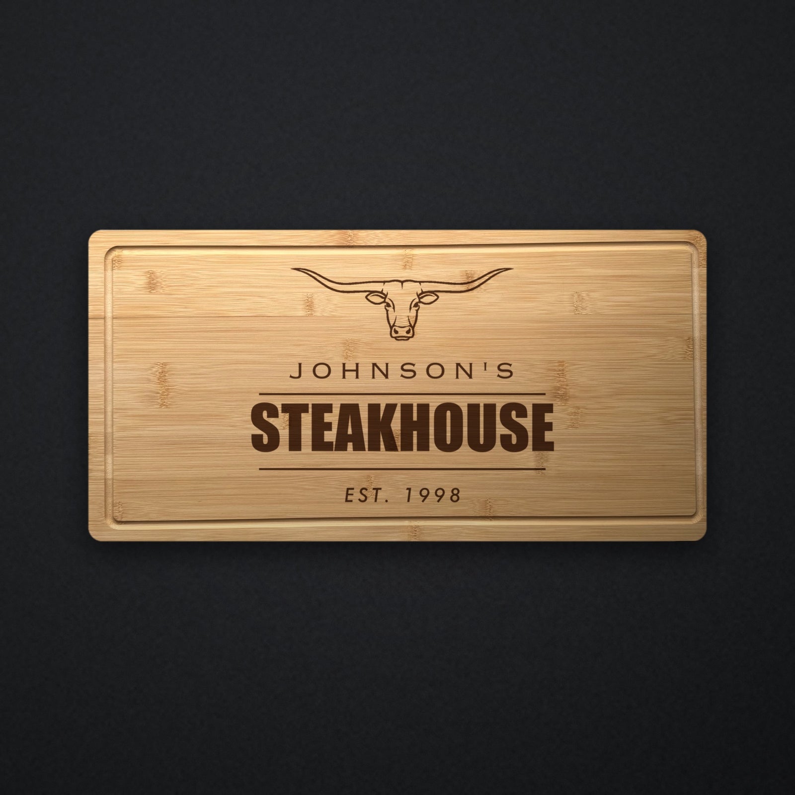 Personalized steakhouse cutting board in solid bamboo on dark slate, engraved with a longhorn steer above the name and the word Steakhouse.
