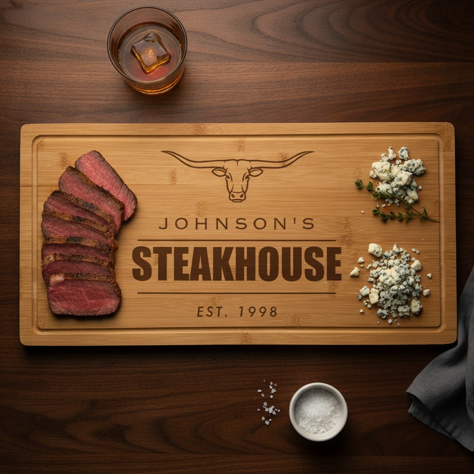 Engraved steakhouse bamboo board on a dark walnut bar top with sliced medium rare ribeye at one end, blue cheese and thyme at the other, beside a bourbon glass.