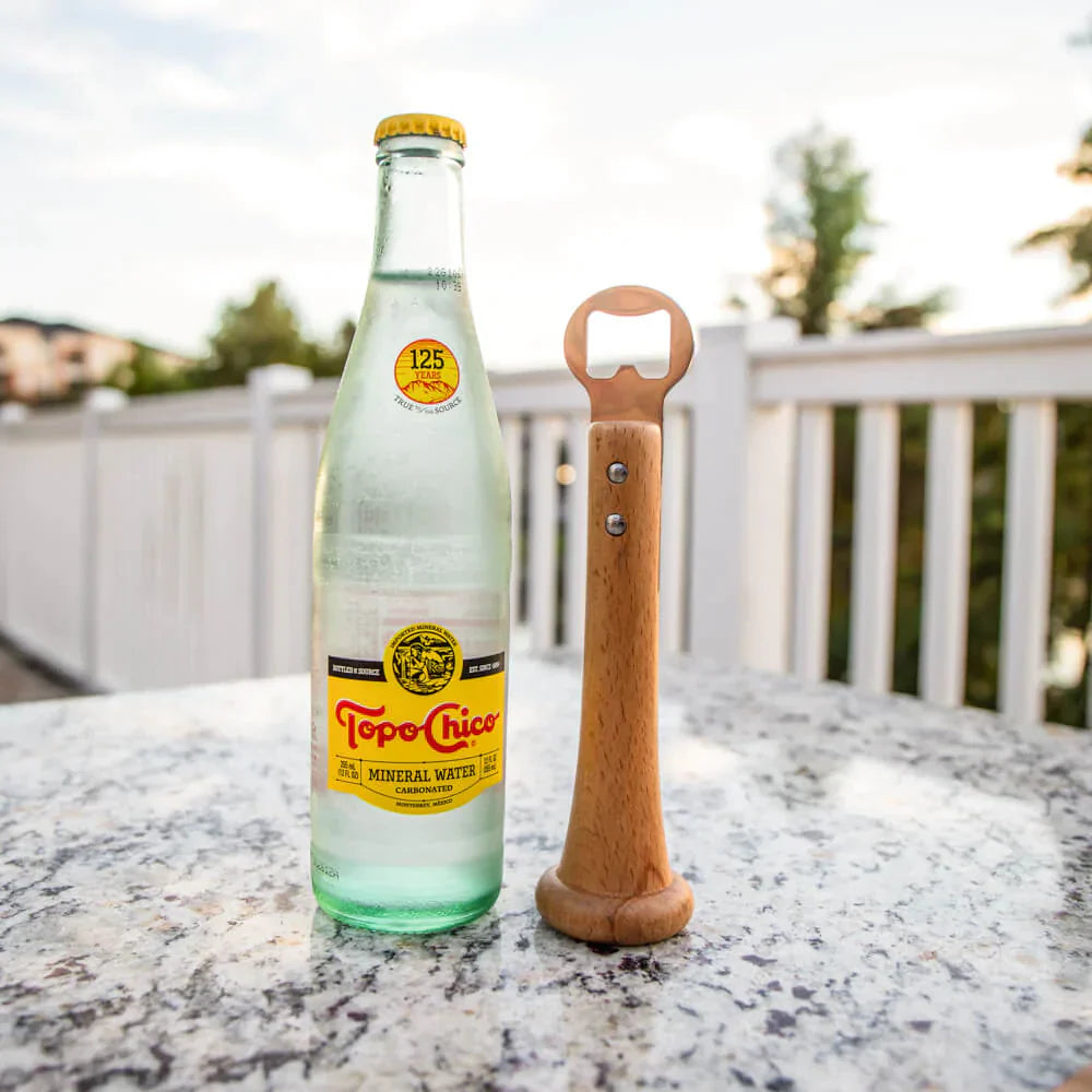 Wooden baseball bat bottle opener displayed next to a beverage bottle on an outdoor stone surface with a deck railing in the background