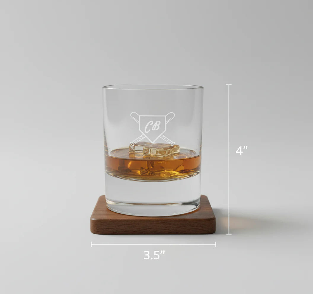 Whiskey glass with engraved sample monogram "CB" and crossed-bats baseball design, filled with amber liquid and ice, displayed on wooden coaster with dimension markers showing 4" height and 3.5" width.