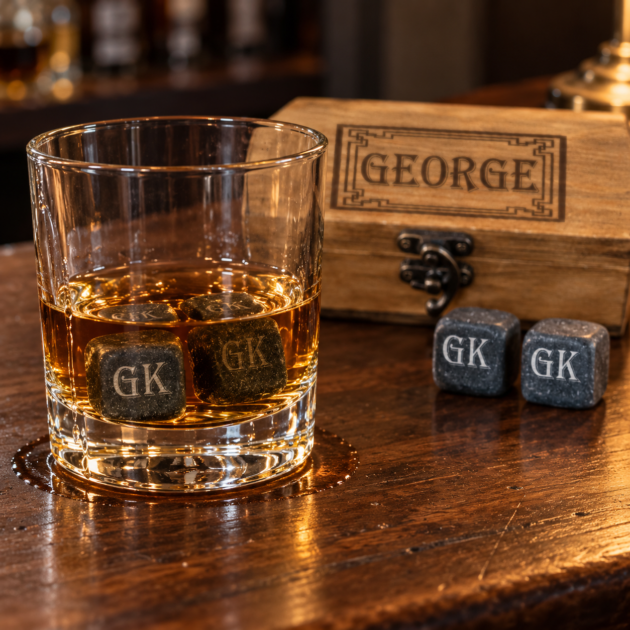 A clear whiskey tumbler with amber liquid and three dark stone dice marked GK, shown on a wooden bar next to a wooden box engraved with GEORGE and two additional GK stones.