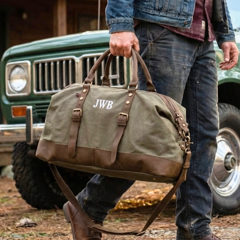 Personalized tan waxed-canvas weekender duffle bag for a groomsman set beside an off-road vehicle, a travel-ready destination wedding gift