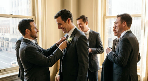 A groom in a charcoal suit with a boutonniere laughs with his four groomsmen in a window-lit suite before the wedding.