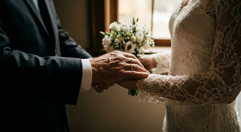 An older father in a dark tuxedo resting his hand on his daughter's lace wedding gown sleeve as she holds her bouquet in the quiet moments before the ceremony