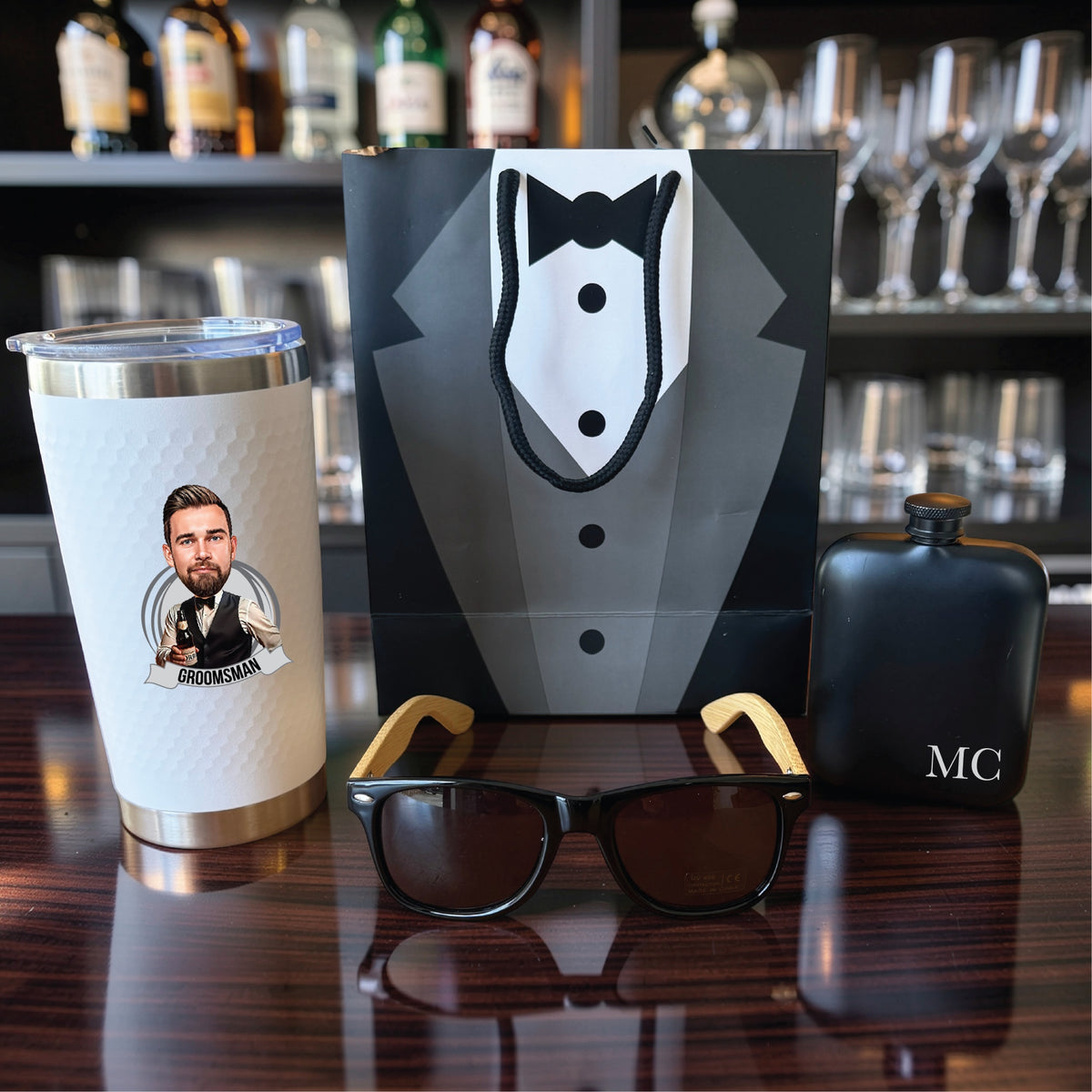 Groomsmen gift set with a caricature tumbler, black flask engraved MC and bamboo sunglasses in front of a tuxedo gift bag on a bar
