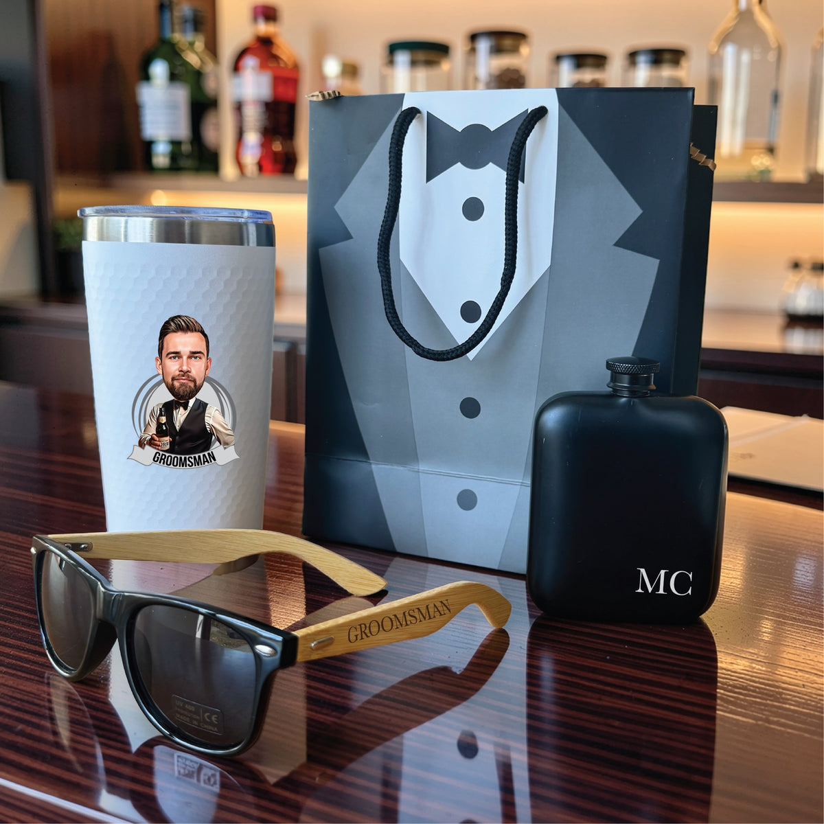 Personalized groomsmen gift set on a bar: tumbler with his caricature, GROOMSMAN engraved sunglasses and an initials flask by a tux gift bag