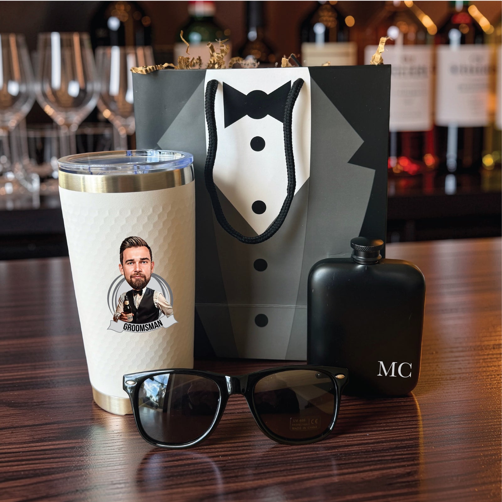 Groomsman caricature tumbler, engraved matte black flask and sunglasses set in front of a tuxedo gift bag with paper shred