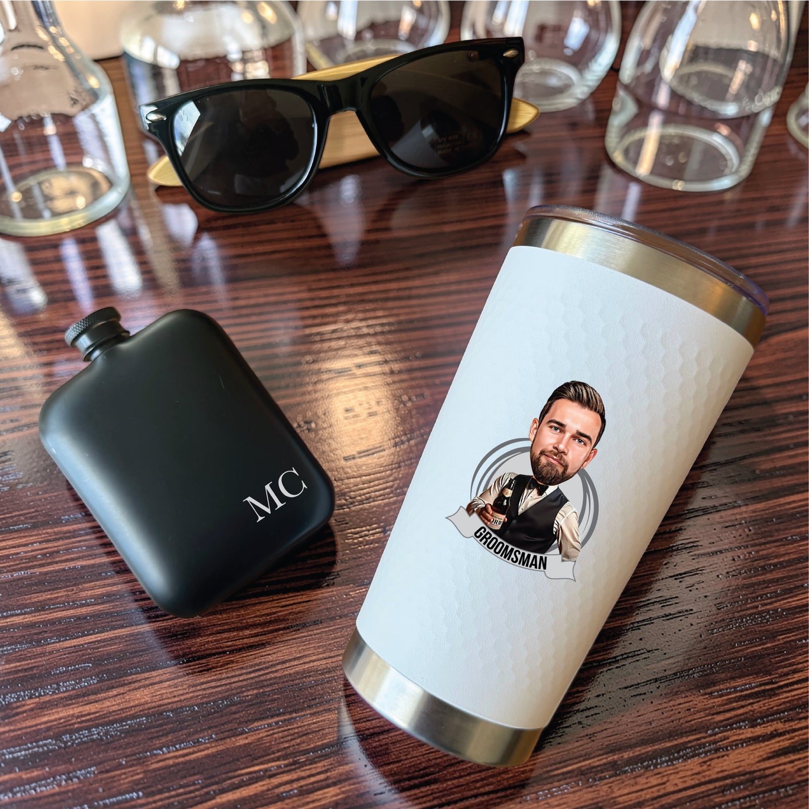 Close-up of the full-color groomsman caricature tumbler beside a matte black flask engraved with the initials MC and bamboo sunglasses
