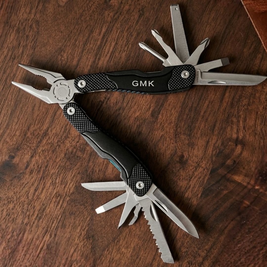 Black multi-tool with carbon fiber handles engraved with sample initials "GMK", displayed open to show multiple stainless steel tools and functions on wooden surface