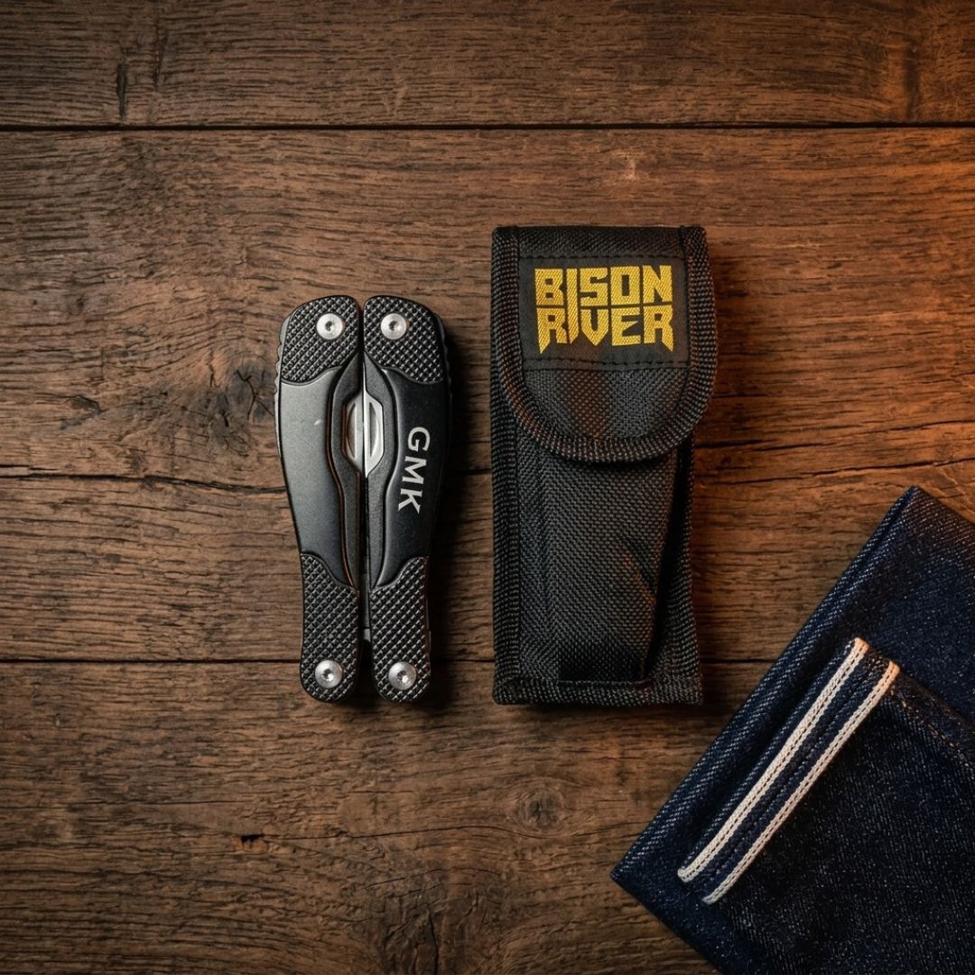 Black multi-tool with textured scales and GMK branding displayed next to black nylon sheath labeled "Bison River" and navy duffel with striped pocket detail on wooden surface.