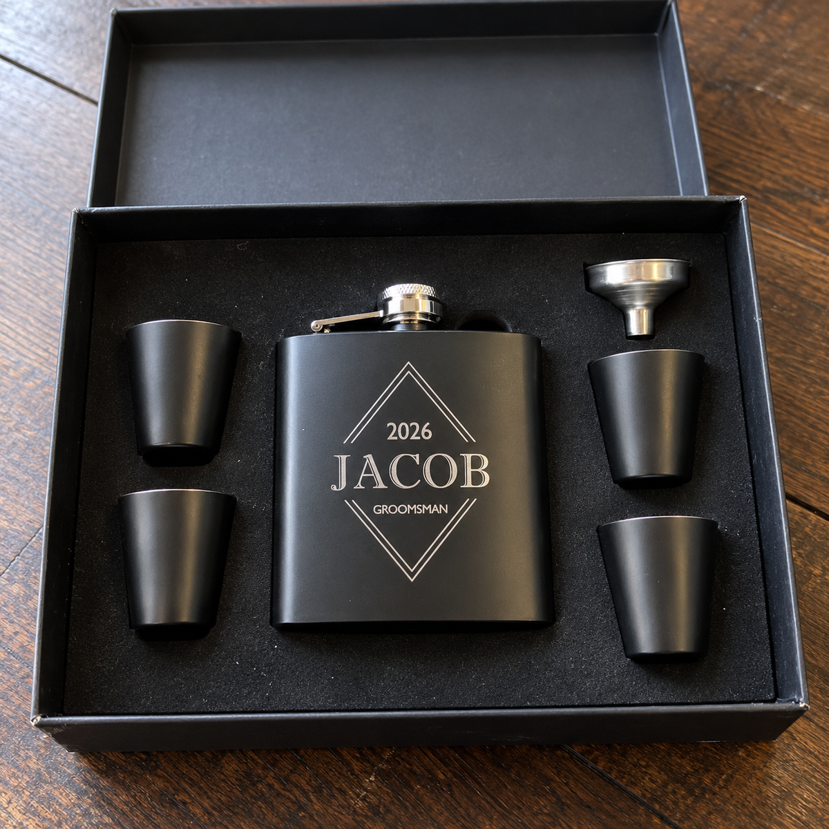 Sharp Shooters groomsmen flask set open in its black gift box, the matte black flask engraved 2026, JACOB and GROOMSMAN beside four shot glasses and a funnel
