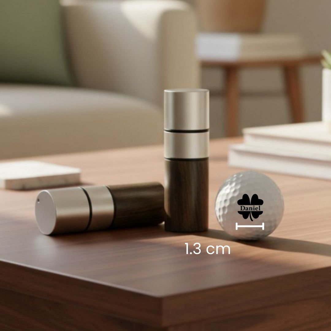 Golf ball stamp with sample personalization showing "Daniel" name and four-leaf clover design, displayed next to stainless steel stamp applicator on wooden table