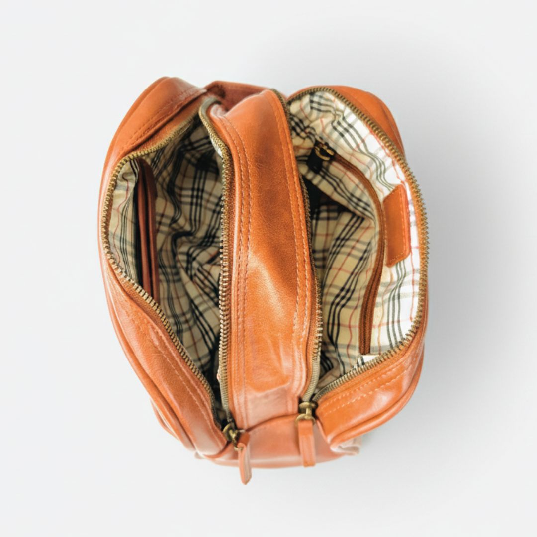 Cognac leather toiletry bag opened flat to display dual compartments and plaid-lined interior with antique silver zippers