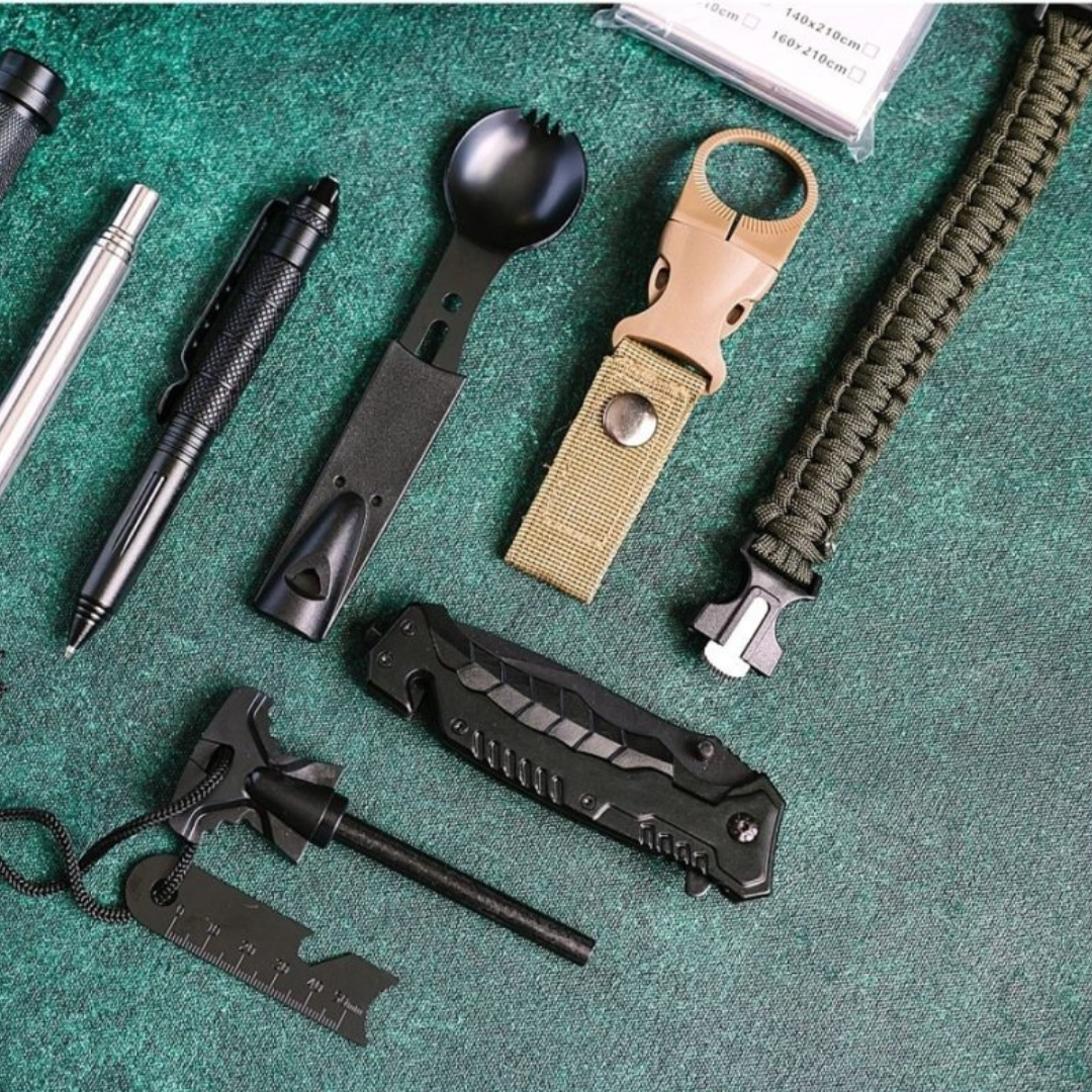 Flat lay of survival kit tools arranged on green surface, including tactical knife, flashlight, fire starter, compass whistle bracelet, carabiner, pen, and additional accessories.