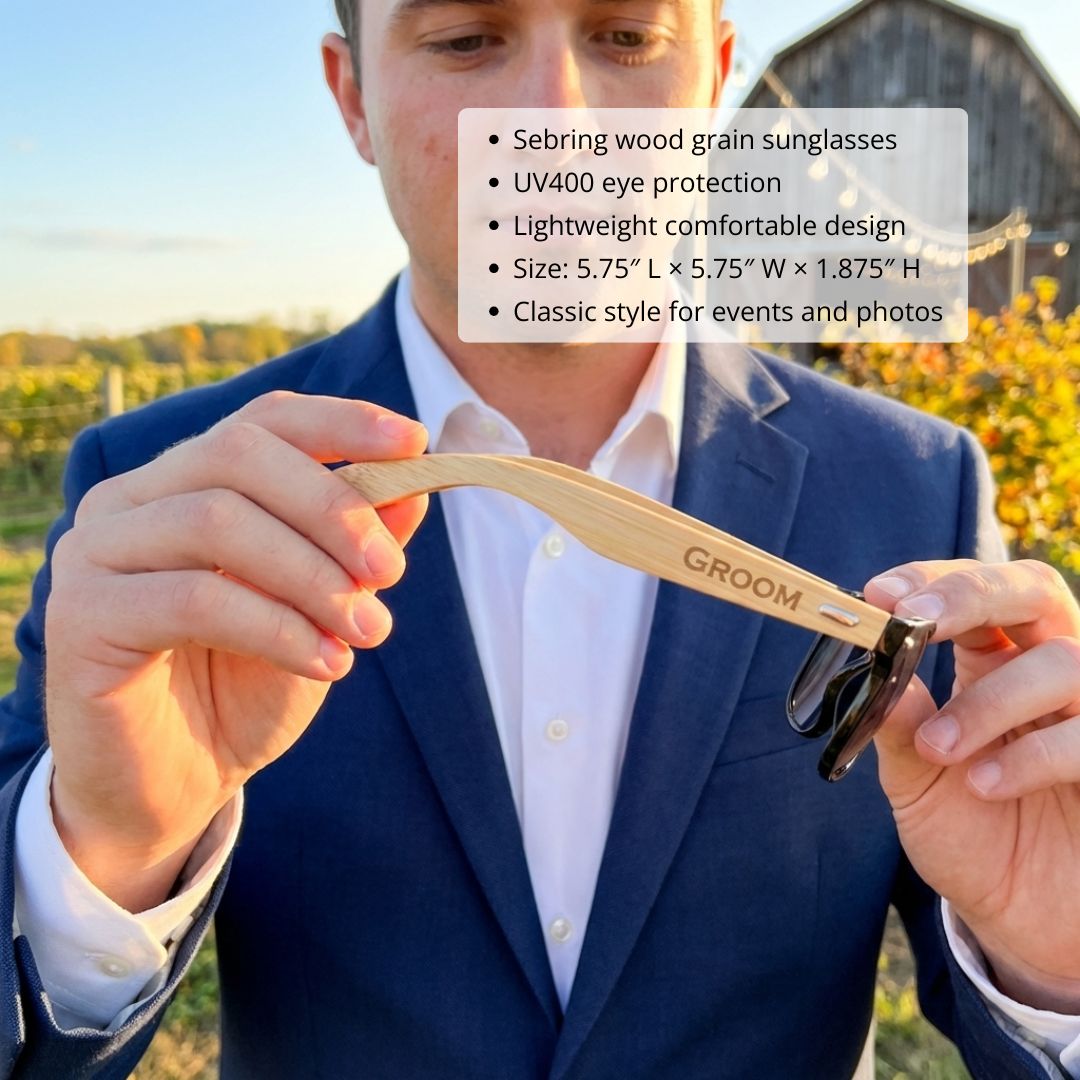 Man in blue suit holding wood-framed sunglasses engraved with sample text "GROOM" on the arm, outdoors at sunset