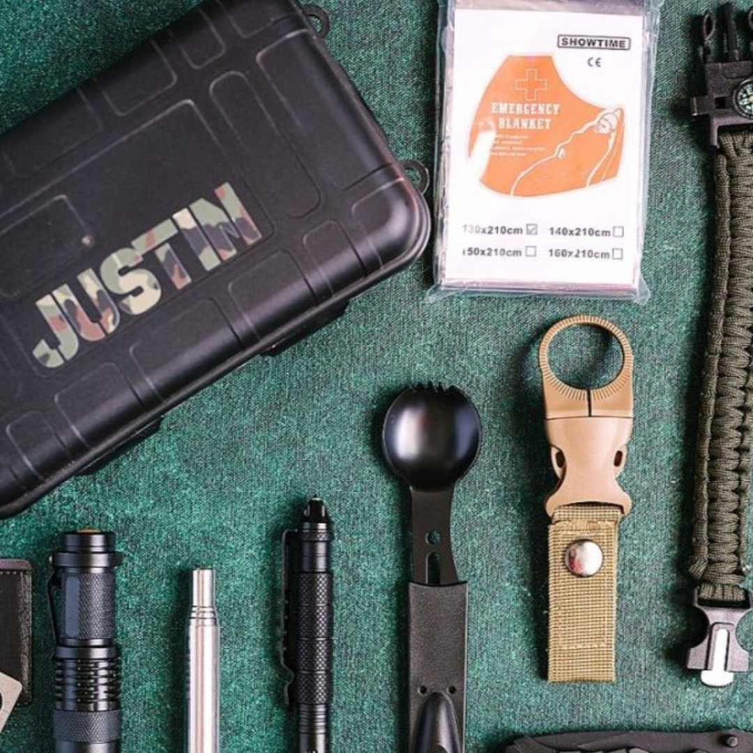 Survival kit contents laid flat on green fabric showing black waterproof case with "JUSTIN" engraving, emergency blanket packaging, flashlight, tactical pen, fire starter, compass bracelet, and additional tools