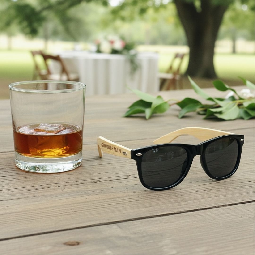 Black frame sunglasses with natural wood temples engraved with sample text "GROOMSMAN," displayed on outdoor wooden table beside whiskey glass.