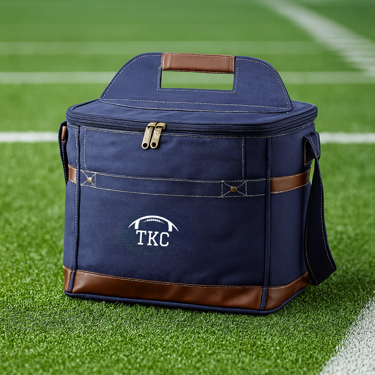 Navy canvas cooler with brown leather trim and handles, embroidered with sample initials "TKC" above a football motif, displayed on artificial turf