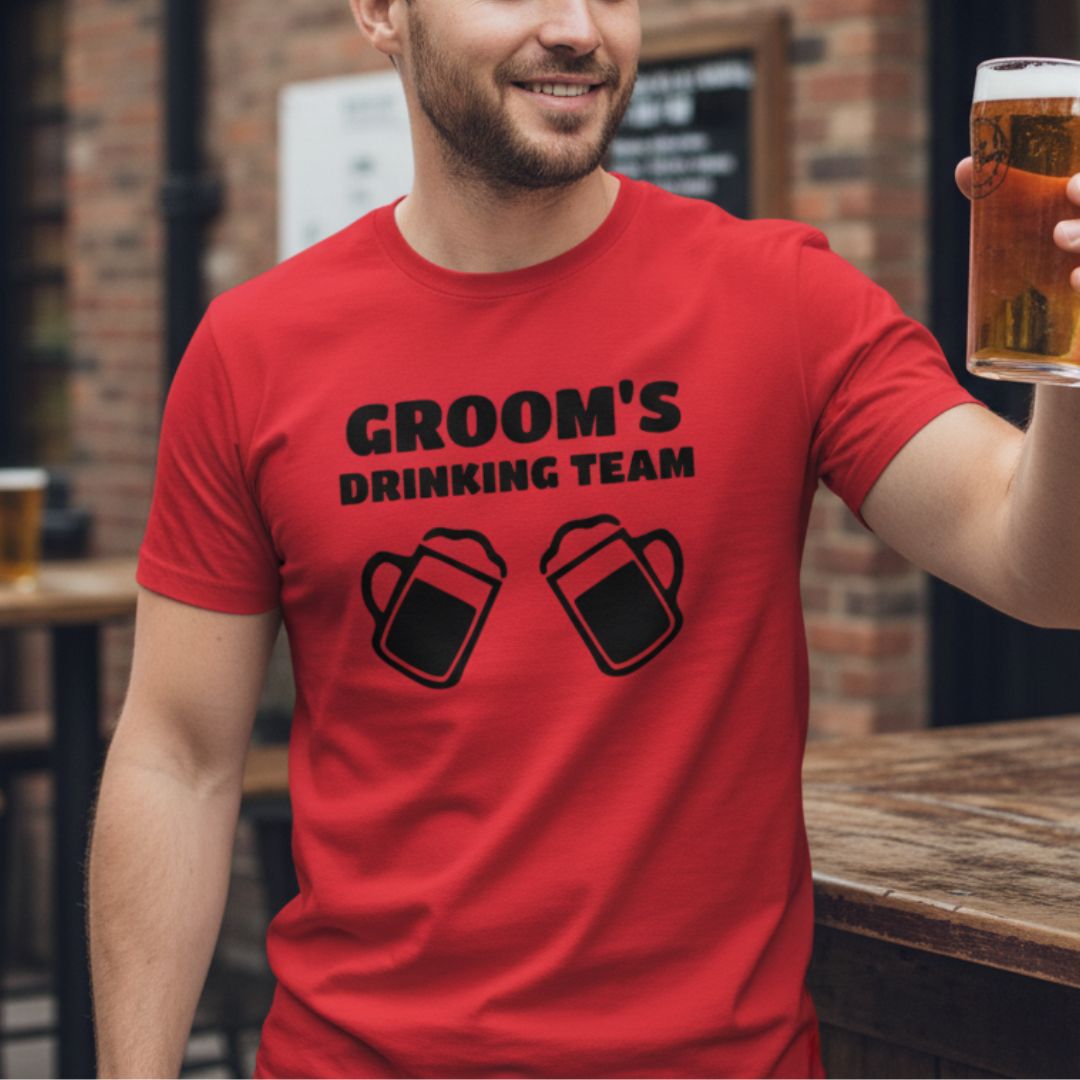 Red t-shirt with black printed text reading "GROOM'S DRINKING TEAM" and two beer mug graphics, modeled by man holding beer glass