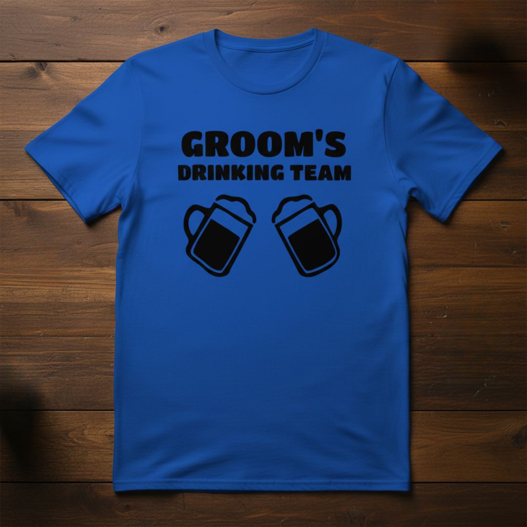 Royal blue t-shirt printed with "GROOM'S DRINKING TEAM" text and two beer mug illustrations in black ink, flat lay on wooden background.