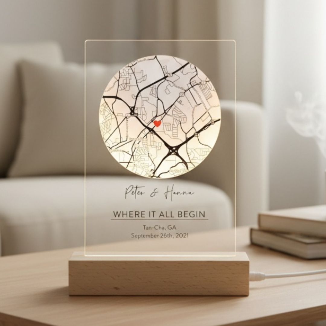 Frosted acrylic sign with circular map showing sample location "Tan-Cha, GA" marked with red heart, personalized with names "Peter &amp; Hanna" and text "WHERE IT ALL BEGAN" with date, mounted on wooden base with warm lighting.