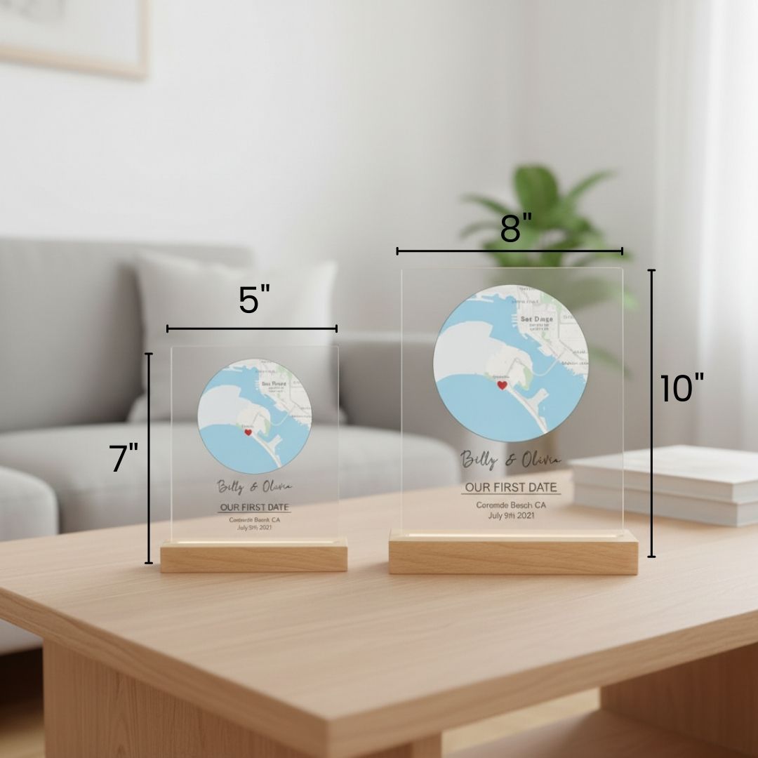 Two acrylic map signs with wood bases displayed on wooden table, smaller 5" by 7" version alongside larger 8" by 10" version, each engraved with sample names "Billy &amp; Olivia", location "Coronado Beach CA", and date "July 9th 2021" with red heart marker.