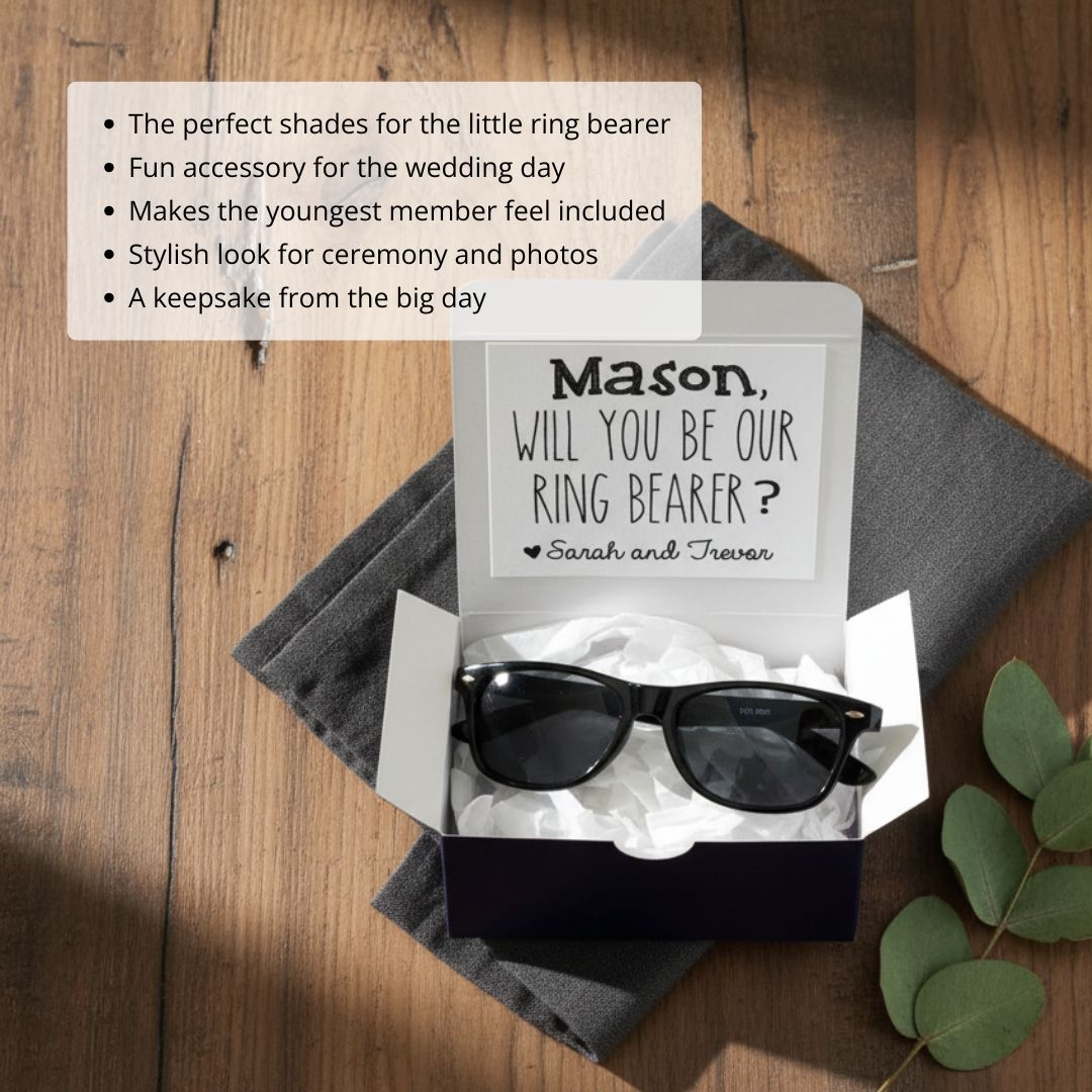 Black wayfarer-style sunglasses displayed in open gift box with personalized message card reading "Will you be our ring bearer?" with sample names and heart detail
