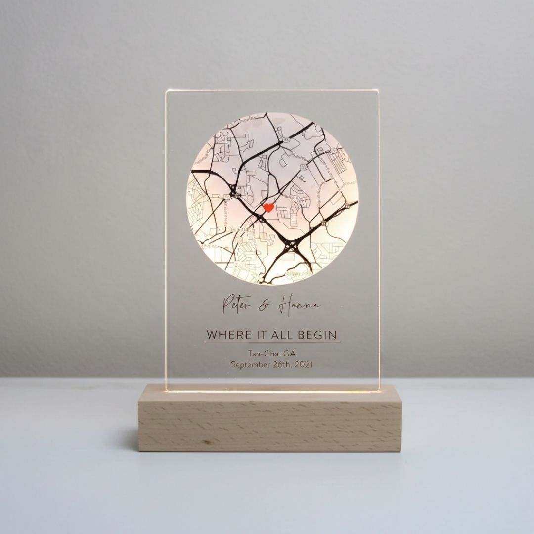 Acrylic map sign with wooden base displaying sample personalization: "Peter &amp; Hanna," "WHERE IT ALL BEGIN," location "Tan-Cha, GA," and date "September 26th, 2021," with circular map showing red heart marker.
