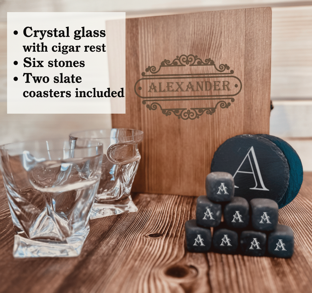 Wooden box engraved with sample name "Alexander" and decorative flourish, displayed with crystal whiskey glasses, slate coasters, and black stones engraved with sample initial "A