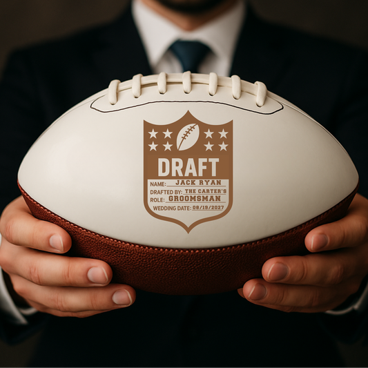 Cream and brown leather football held in hands, engraved with sample "Draft" shield design featuring name "Jack Ryan," role "Groomsman," and wedding date "08/19/2027