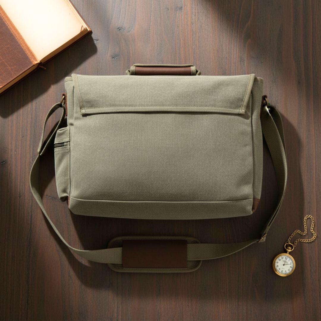 Sage canvas messenger bag with brown leather handles and trim, displayed flat on wooden surface with pocket detail visible from back.