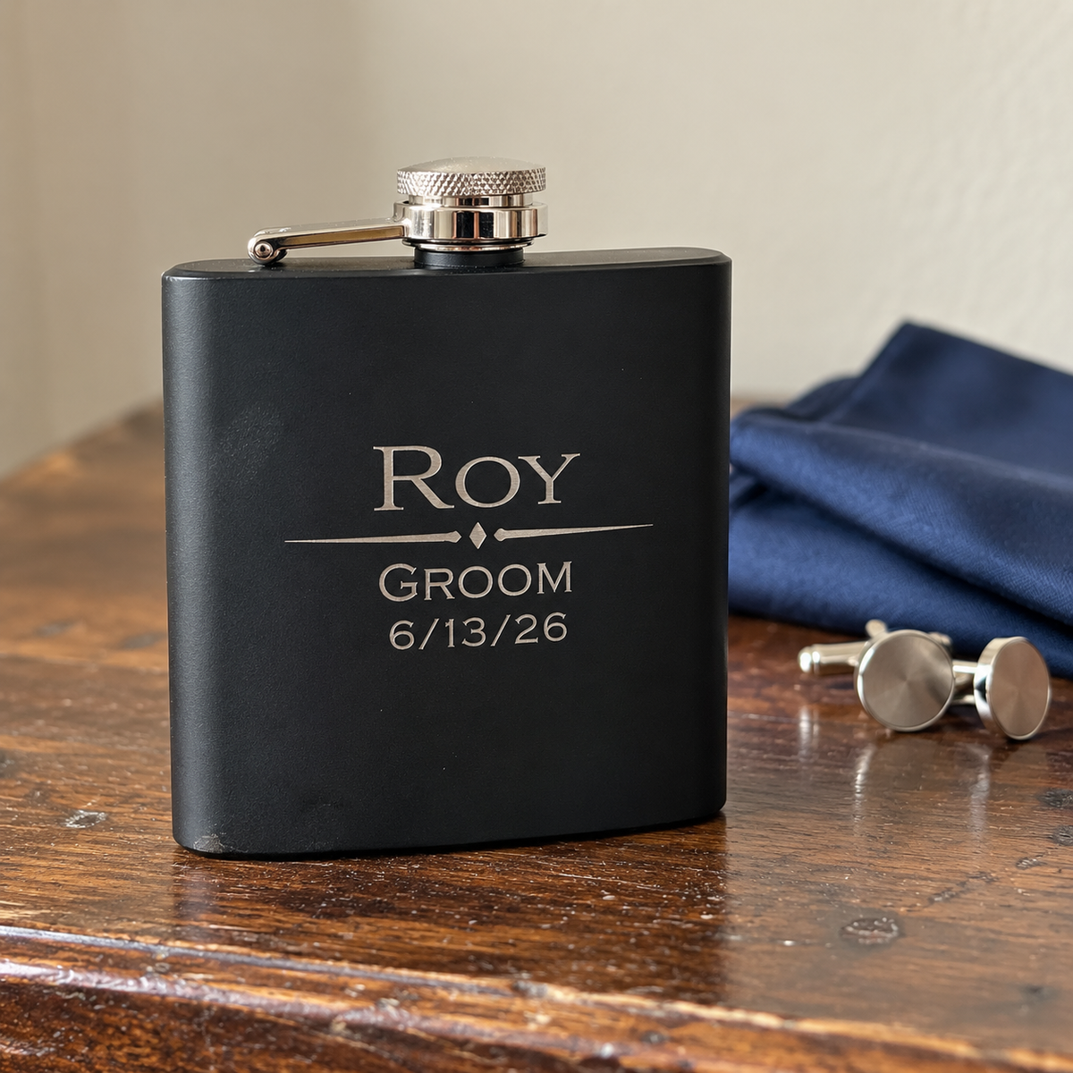 Tin Diesel matte black groomsmen flask engraved Roy, Groom and 6/13/26 in light silver on the front, standing on a walnut dresser beside cufflinks