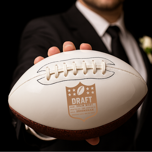 White leather football with tan laces held in hands, engraved with sample "Draft" label showing name Colin West, role Groomsman, drafting team The Carter's, and wedding date 09/15/2027