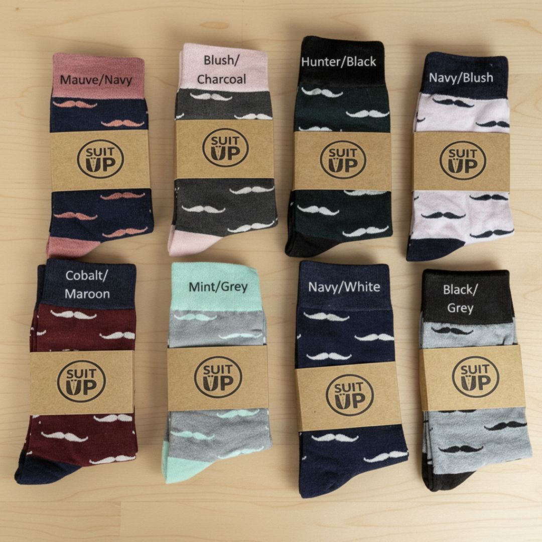 Eight pairs of socks arranged in a grid, each displayed with kraft paper band labels reading "SUIT UP." Socks feature white mustache patterns on various color combinations including mauve/navy, blush/charcoal, hunter/black, navy/blush, cobalt/maroon, mint/grey, navy/white, and black/grey colorways.