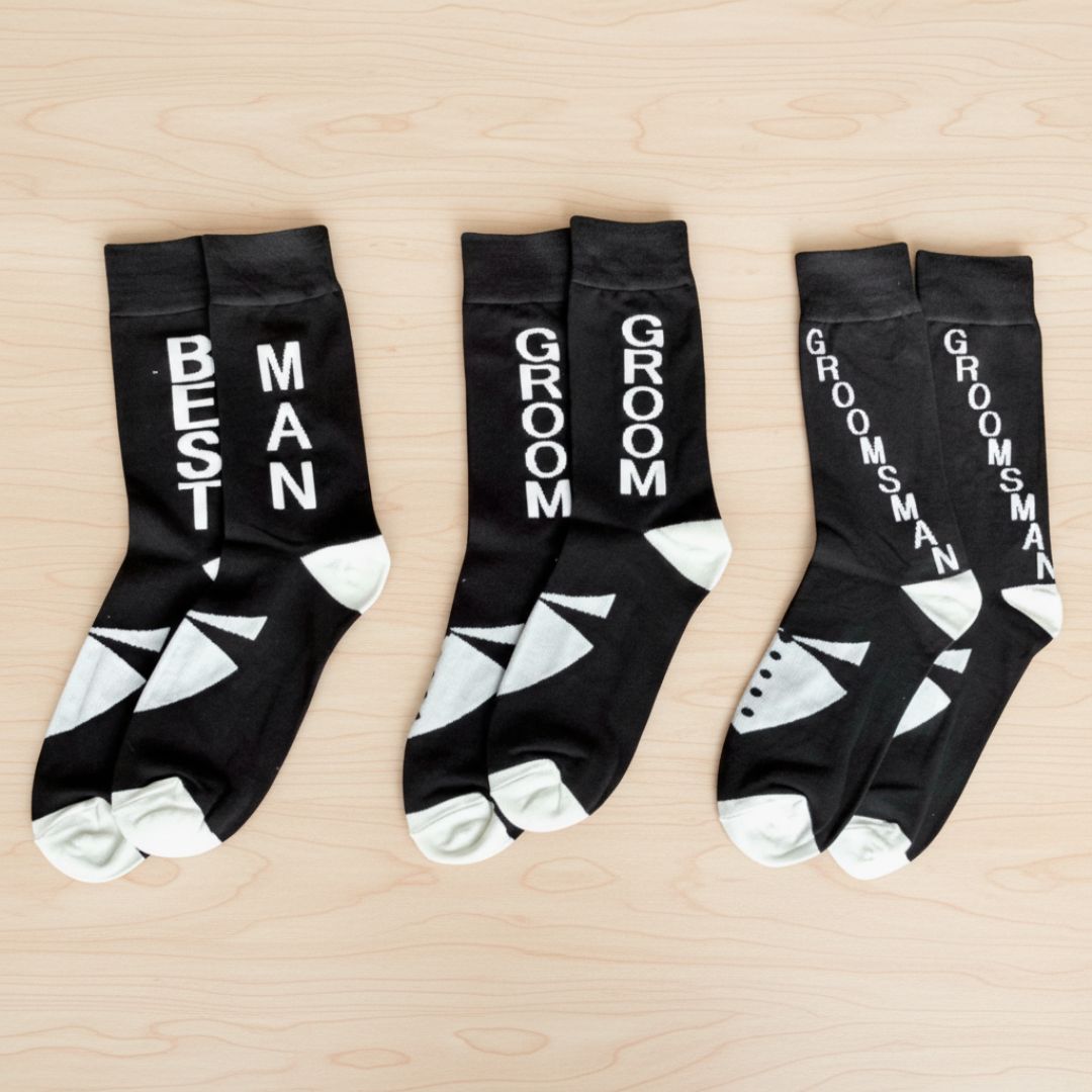 Three black crew socks with white text and toe panels displayed flat, showing sample designs reading "Best Man," "Groom," and "Groomsman" on each pair