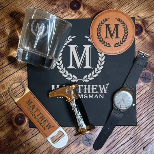Personalized groomsmen gift box pieces on wood: laurel monogram rocks glass, tan coaster, engraved bottle opener, gold torch lighter and wood watch